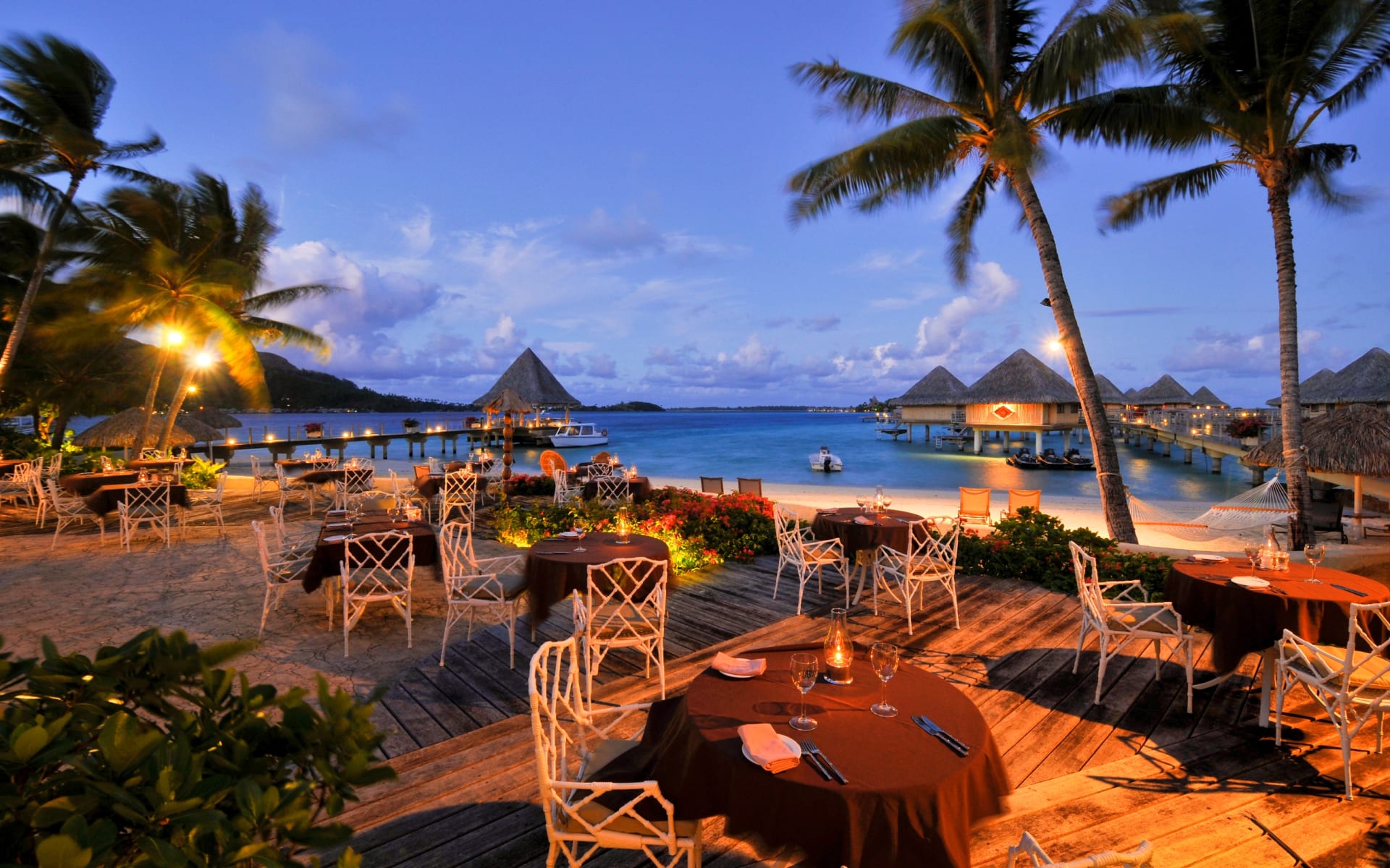 It's nighttime, and the dining tables overlook the beach and are backed by palm trees and lamps. 